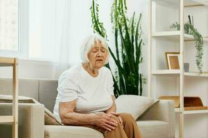 ouderen vrouw erge, ernstige pijn in haar been zittend Aan de bank, Gezondheid problemen in oud leeftijd, arm kwaliteit van leven. grootmoeder met grijs haar- houdt Aan naar haar zeer knie, problemen met gewrichten en ligamenten foto