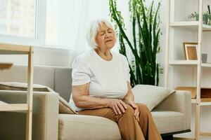 ouderen vrouw erge, ernstige pijn in haar been zittend Aan de bank, Gezondheid problemen in oud leeftijd, arm kwaliteit van leven. grootmoeder met grijs haar- houdt Aan naar haar zeer knie, problemen met gewrichten en ligamenten foto