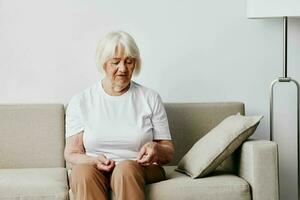 ouderen vrouw zit Aan sofa Bij huis, helder ruim interieur in oud leeftijd glimlach, levensstijl. grootmoeder met grijs haar- in een wit t-shirt en beige broek. foto