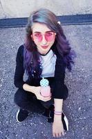 gelukkige mooie tiener met roze zonnebril drinkt en geniet van een roze drankje zittend op stedelijke grond foto