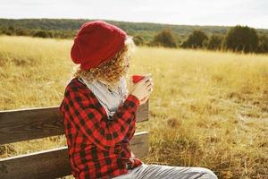 een jonge vrouw van achteren in een rood geruit hemd met een wollen muts en sjaal die een kopje thee of koffie neemt terwijl ze zonnebaadt zittend op een houten bank in een geel veld met achtergrondverlichting van de herfstzon foto
