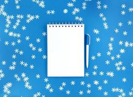 wit blanco vel notebook en een pen op een blauwe achtergrond met verspreide confetti sneeuwvlokken. vakantie onderwijs concept. Stock foto.