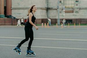 atletisch vrouw Aan rol skates beweegt actief poses Aan asfalt Bij straat gekleed in actief slijtage heeft vrolijk uitdrukking. jong geschiktheid model- het schaatsen in stedelijk plaats. sportief levensstijl skaten foto