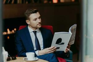 aantrekkelijk zakenman in pak, leest tijdschrift, drankjes cappuccino, wacht voor partner, gefocust. mensen en rust uit foto