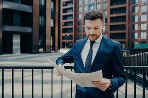 gefocust zakenman in pak cheques krant- voor interessant nieuws, staat alleen buitenshuis, leunend Aan zwart hek door kantoor plein gebouwen, genieten van werk pauze. foto