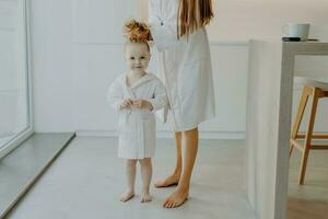 schattig meisje in wit gewaad poses met mama, haar- gekamd, paardenstaart, kaal voeten. haarverzorging en kinderen concept. foto