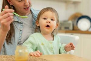 gelukkig familie Bij huis. moeder voeden haar baby meisje van lepel in keuken. weinig kleuter kind met rommelig grappig gezicht eet gezond voedsel Bij huis. jong vrouw mam geven voedsel naar kind dochter. foto