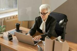 zelfverzekerd elegant Europese midden- oud senior vrouw gebruik makend van laptop Bij werkplek. elegant ouder volwassen Jaren 60 grijs haren dame zakenvrouw zittend Bij kantoor tafel. baas leider leraar professioneel arbeider. foto