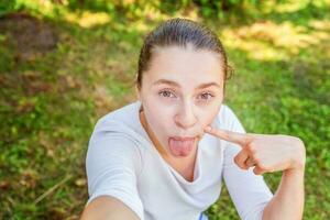 jong meisje nemen selfie van handen met telefoon tonen tong en grappig gezicht zittend Aan park of tuin achtergrond foto