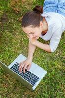jong vrouw aan het liegen Aan groen gras gazon in stad park werken Aan laptop pc computer. freelance bedrijf concept foto
