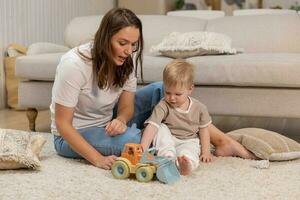 gelukkig familie Bij huis. moeder en baby jongen spelen met speelgoed Bij huis binnenshuis. weinig kleuter kind en oppas oppas hebben pret samen. jong vrouw mam kind zoon rust uit in leven kamer. foto