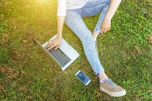 vrouw benen op groen gras gazon in stadspark, handen werken op laptop pc-computer. freelance bedrijfsconcept foto