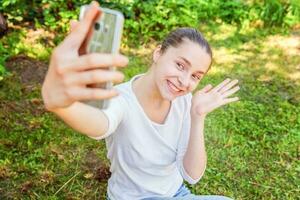jong grappig meisje nemen selfie van handen met telefoon zittend Aan groen gras park of tuin achtergrond foto