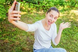 jong grappig meisje nemen selfie van handen met telefoon zittend Aan groen gras park of tuin achtergrond foto