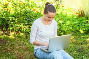 jonge vrouw zittend op groen gras gazon in stadspark werken op laptop pc-computer. freelance bedrijfsconcept foto