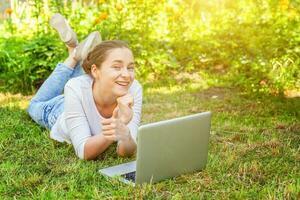 jong vrouw aan het liegen Aan groen gras gazon in stad park werken Aan laptop pc computer. freelance bedrijf concept foto