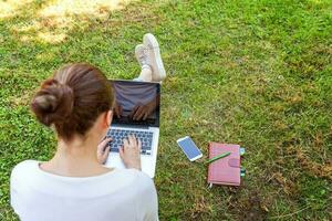 jonge vrouw zittend op groen gras gazon in stadspark werken op laptop pc-computer. freelance bedrijfsconcept foto