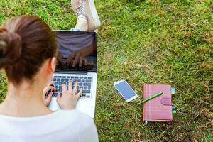 jonge vrouw zittend op groen gras gazon in stadspark werken op laptop pc-computer. freelance bedrijfsconcept foto