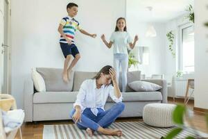 moe jong moeder zittend Aan de verdieping en werken met laptop en documenten terwijl weinig kinderen en jumping Aan de sofa en hebben pret en maken lawaai foto