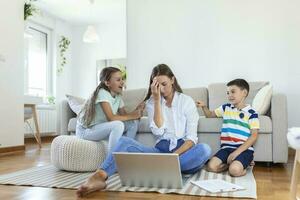 klein ondeugend kinderen lawaai maken en afleidend moeder freelancer proberen naar concentreren Aan laptop en knijpen hoofd werken Bij tafel in licht leven kamer foto