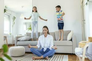 jong moeder met Gesloten ogen mediteren in lotus houding Aan verdieping proberen naar opslaan binnenste harmonie terwijl opgewonden kinderen jumping Aan sofa en schreeuwen in licht ruim leven roo foto