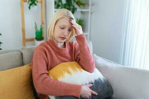 portret van een jong blond vrouw zittend Aan de bankstel Bij huis met een hoofdpijn en migraine . mooi vrouw lijden van chronisch dagelijks hoofdpijn. verdrietig vrouw Holding haar hoofd omdat sinus pijn foto