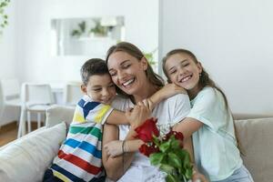 jong moeder met een boeket van rozen lacht, knuffelen haar zoon, en vrolijk meisje met een kaart feliciteert mam gedurende vakantie viering in keuken Bij huis foto