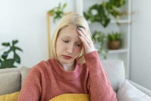 portret van een jong blond vrouw zittend Aan de bankstel Bij huis met een hoofdpijn en migraine . mooi vrouw lijden van chronisch dagelijks hoofdpijn. verdrietig vrouw Holding haar hoofd omdat sinus pijn foto