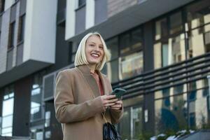 prachtig mooi jong vrouw met blond haar- berichten Aan de smartphone Bij de stad straat achtergrond. mooi meisje hebben slim telefoon gesprek foto