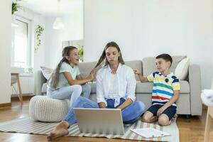 klein ondeugend kinderen lawaai maken en afleidend moeder freelancer proberen naar concentreren Aan laptop en knijpen hoofd werken Bij tafel in licht leven kamer foto