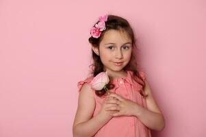 authentiek Kaukasisch mooi elegant weinig meisje in roze jurk, Holding een roos bloem, glimlachen schattig op zoek Bij camera foto