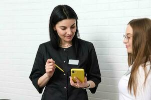 vrouw dokter, tandarts of schoonheidsspecialist in zwart uniform, cheques haar telefoon, staat in de buurt medisch assistent Bij ontvangst bureau foto