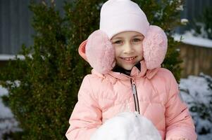 Kaukasisch weinig kind meisje in warm winter kleren en roze pluizig oorwarmers, glimlacht Bij camera, gebouw sneeuwman buitenshuis foto