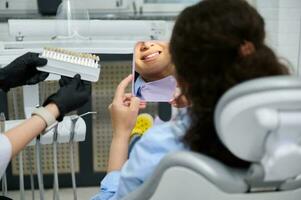 achterzijde visie vrouw in tandheelkundig stoel, glimlacht Bij spiegel, tandarts gebruik makend van tand schaduw grafiek, met vergelijking fineer- schaal tabel foto
