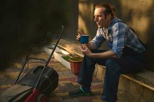 professioneel tuinman ontspant met een mok van koffie in de buurt een gazon maaier in de tuin na moeilijk dag werk. mensen arbeid foto