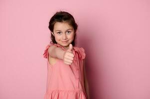 Kaukasisch aanbiddelijk weinig kind meisje in roze jurk, duimen omhoog en glimlachen op zoek Bij camera, geïsoleerd Aan roze achtergrond foto