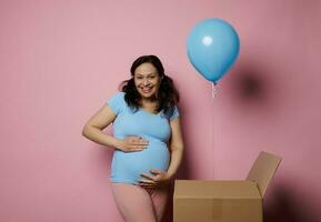 emotioneel charismatisch zwanger vrouw aanraken haar buik, glimlachen Bij camera, geïsoleerd roze achtergrond met blauw ballon. foto