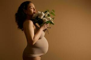 aantrekkelijk etnisch zwanger vrouw, verwachtend moeder met een bundel van seringen, poseren in beige lingerie. studio portret foto