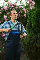 professioneel mannetje tuinman in blauw uniform, gebruik makend van snoeiers, snijdend een haag in binnenplaats van een herenhuis. tuin decoratie foto