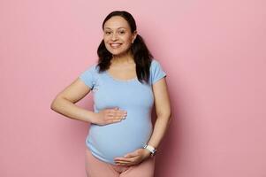 charmant zwanger gravid vrouw met armen Aan haar ronde buik, glimlachen Bij camera, geïsoleerd roze achtergrond. ervan uitgaand baby foto