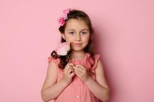 detailopname weinig kind meisje in roze jurk en bloemen in kapsel, Holding een roze roos bloem, lachend, geïsoleerd Aan roze foto