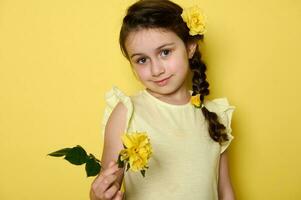 schoonheid portret van een Kaukasisch edele charmant baby meisje in geel jurk en bloemen in kapsel, Holding geel roos. foto