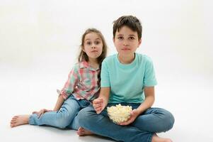 aanbiddelijk kinderen, pre tiener jongen en weinig meisje aan het eten popcorn, aan het kijken film of tekenfilm, wit achtergrond. familie vrije tijd foto