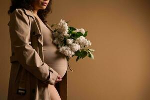 detailopname zwanger vrouw met een bundel van vers bloeiend seringen, Holding haar buik, geïsoleerd over- beige studio achtergrond foto