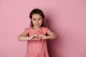 mooi blauw ogen weinig meisje in roze jurk, vormen hart vorm met handen, glimlachen Bij camera, geïsoleerd roze backdrop foto