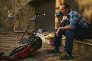 professioneel tuinman in blauw tuinieren jumpsuit en plaid shirt, nemen een breken na moeilijk werken dag in de tuin foto