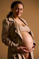 mooi zwanger vrouw in beige loopgraaf jas, voorzichtig strelen haar kaal buik, op zoek Bij camera, geïsoleerd Aan beige foto