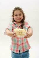 gelukkig vrolijk weinig meisje Holding uit Bij camera een kom met gezouten popcorn, glimlachen breed Aan wit geïsoleerd achtergrond foto