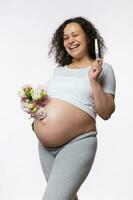 glimlachen gelukkig zwanger vrouw tonen Bij camera een positief zwangerschap testen, poseren met bundel van bloemen. menselijk vruchtbaarheid. foto