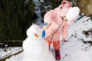 lief weinig kind meisje in roze naar beneden jasje en pluizig oorwarmers Holding een Schep en gebouw een sneeuwman buitenshuis foto